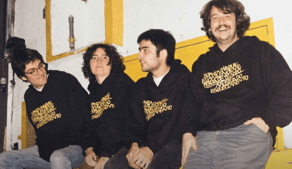 Da esquerda para a direita: Téo Serson, Sophia, Vicente e Theo Ceccato, sentados lado a lado em um ambiente interno com parede branca e detalhes amarelos. Todas vestem moletons pretos com estampa em letras amarelas. Duas delas olham para frente, uma olha para o lado e outra sorri para a câmera.