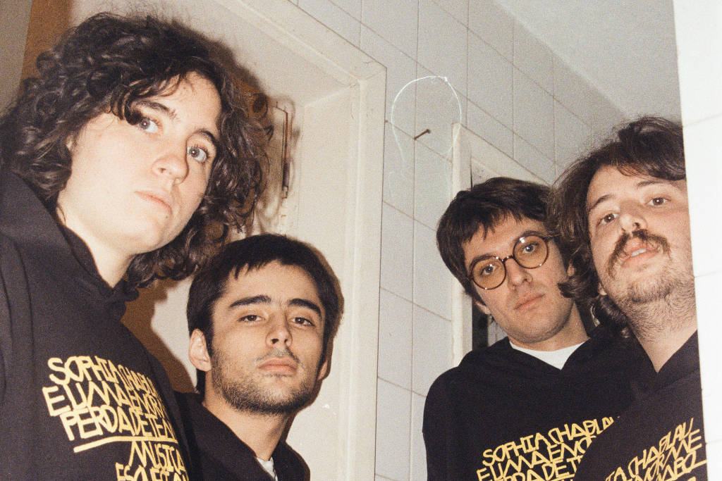 Quatro pessoas jovens posando juntas em um ambiente interno, próximo a uma porta e uma parede de azulejos brancos. Todas vestem moletons pretos com uma estampa em letras amarelas. Três delas olham diretamente para a câmera, enquanto uma olha levemente de lado.