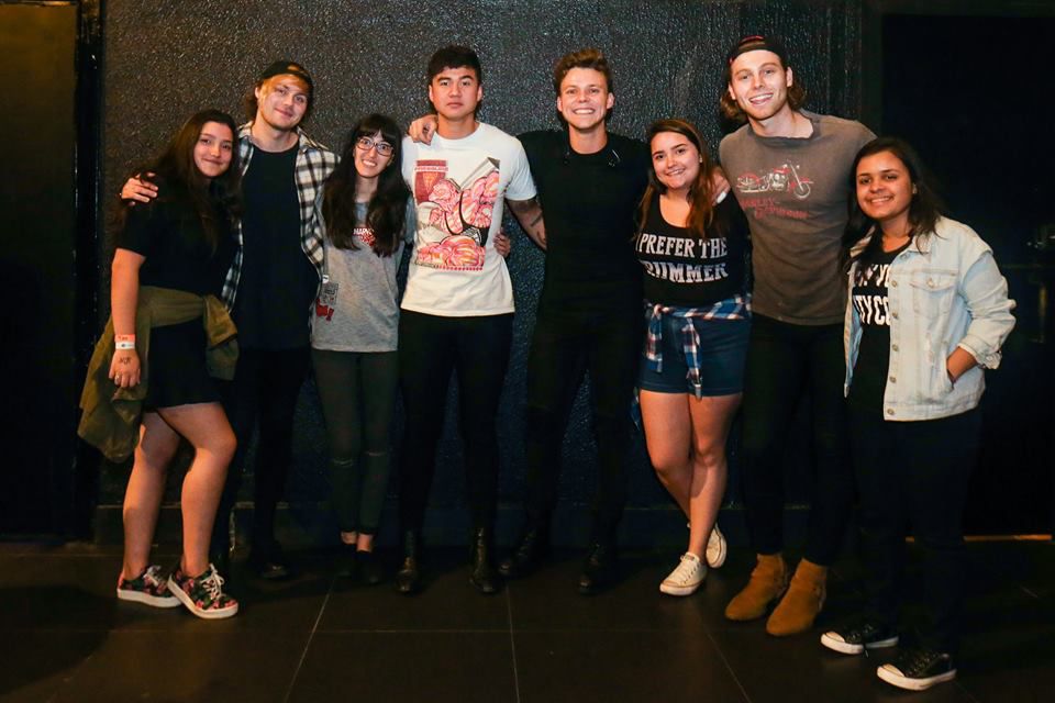 Um grupo de nove pessoas posa lado a lado em frente a uma parede escura, com quatro homens ao centro e cinco mulheres ao redor, todos sorrindo. O clima é casual e parece um meet & greet.