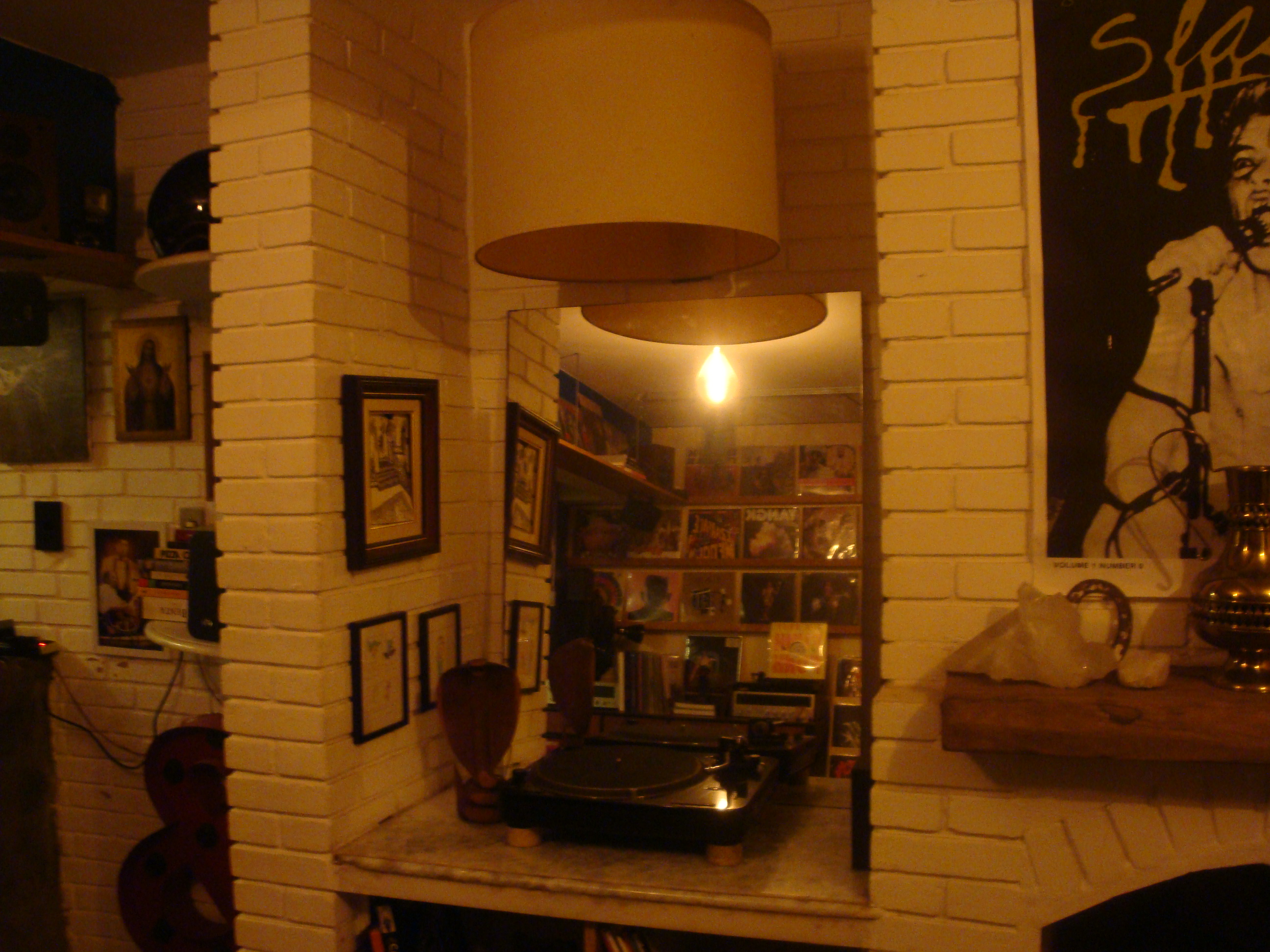 Canto de uma sala com parede branca de tijolinhos, toca-discos sobre uma bancada e um espelho refletindo mais vinis.